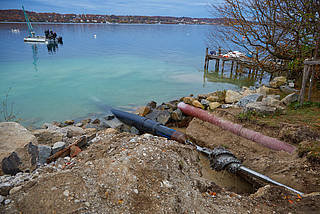 Die HDD-Bohrung der Seeleitung im Starnberger See wird erstellt und eingezogen. Rechts ist ein Erdloch mit Bohrwelle und Rohren zu sehen. Im Hintergrund der Starnberger See.
