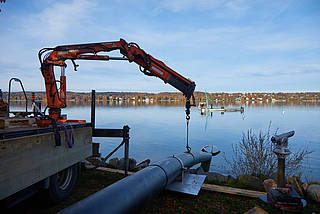 Ein Greifarm befördert die Rohrleitung hinunter in den Starnberger See. Im Hintergrund ist das andere Ufer mit Wald und Häusern zu erkennen.
