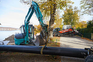 Ein grüner Bagger steht vor der Rohrleitung und hebt diese mithilfe eines Gurtes an. Im Hintergrund ist herbstliches Laub und weitere Maschinen erkennbar.